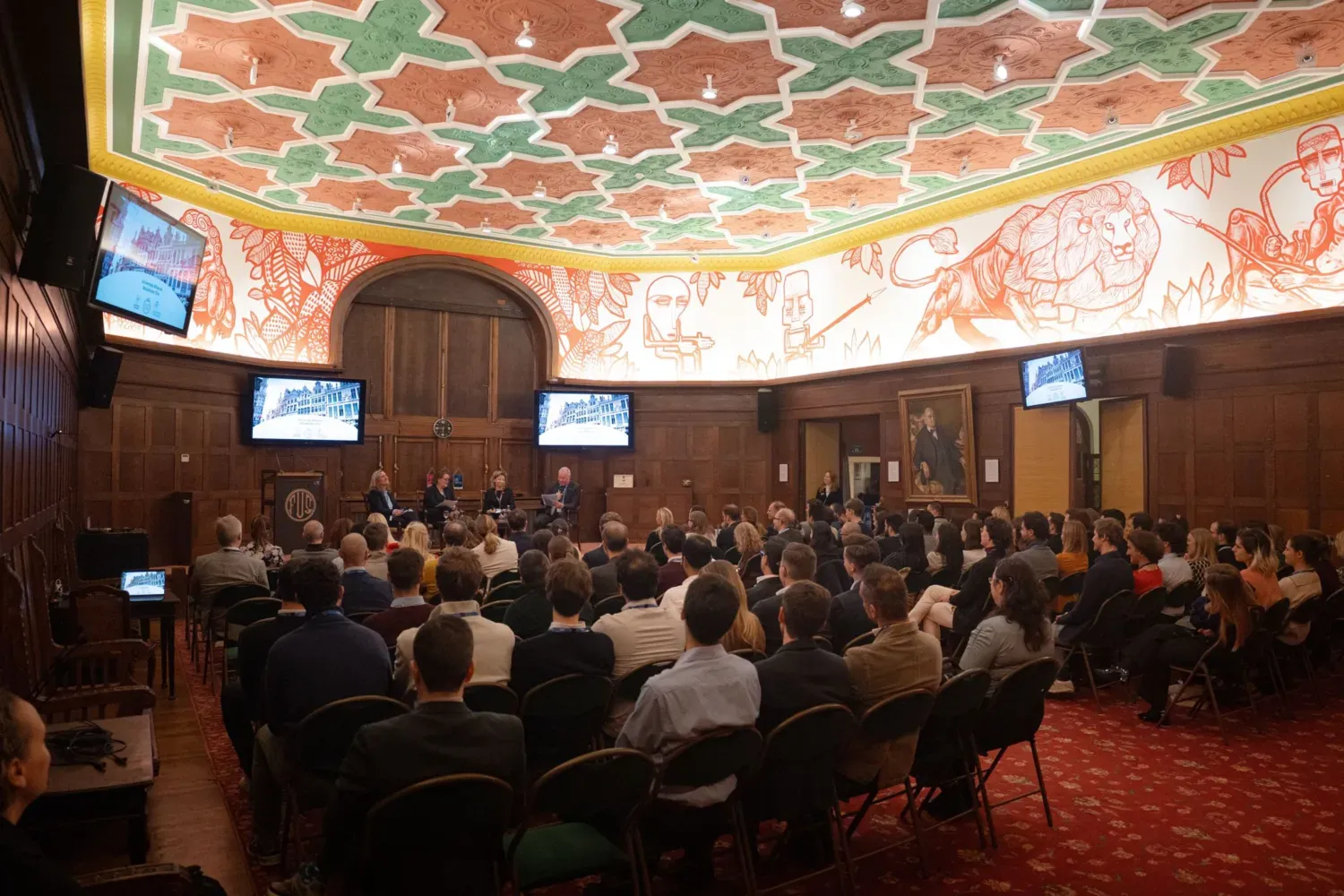An audience is seated in a grand hall, facing a stage, where four people are seated with mics.
