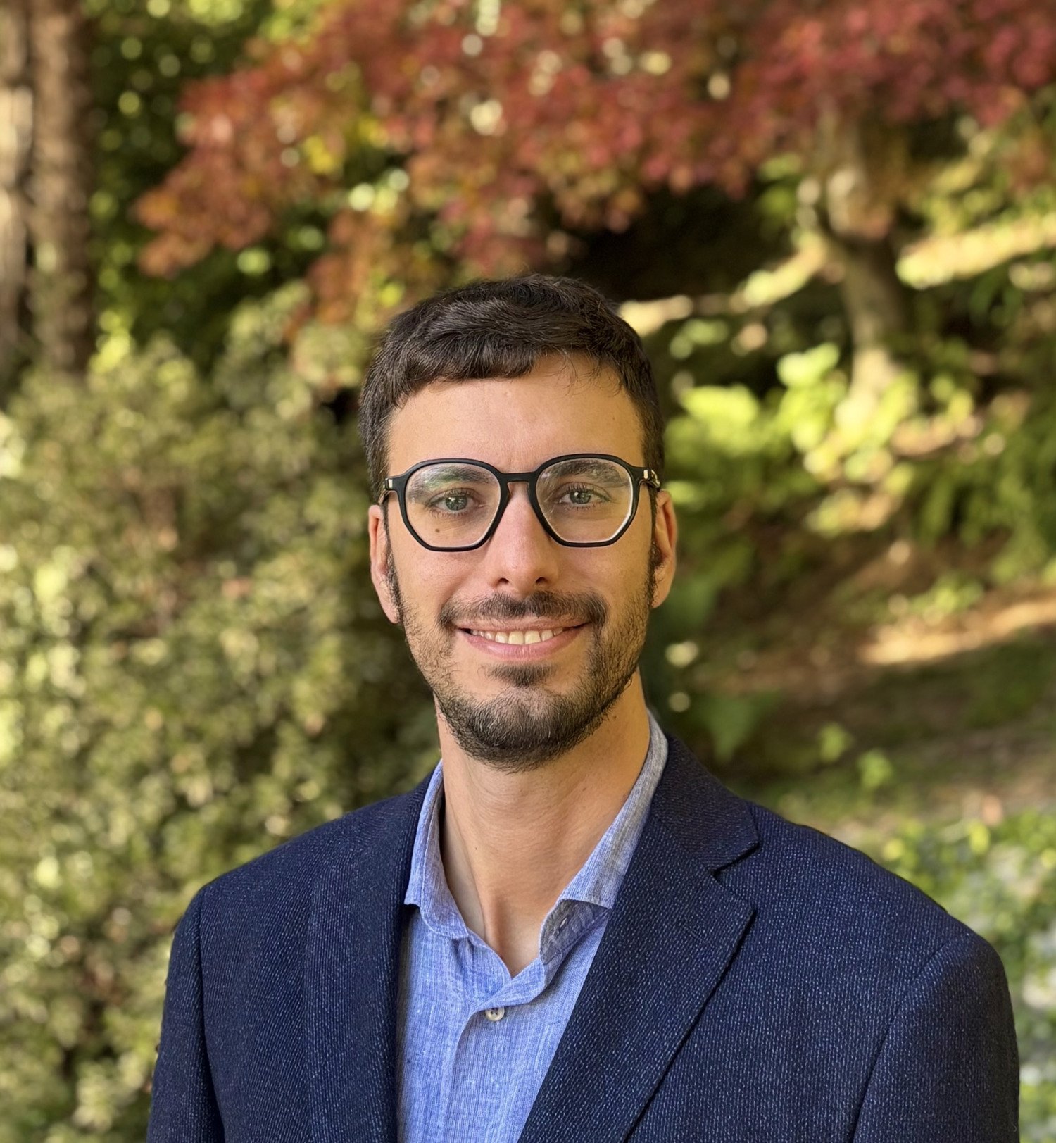 Portrait photo of a man. He is wearing glasses, a blue shirt and a blue jacket. In the background, there is a green lawn, trees and bushes.