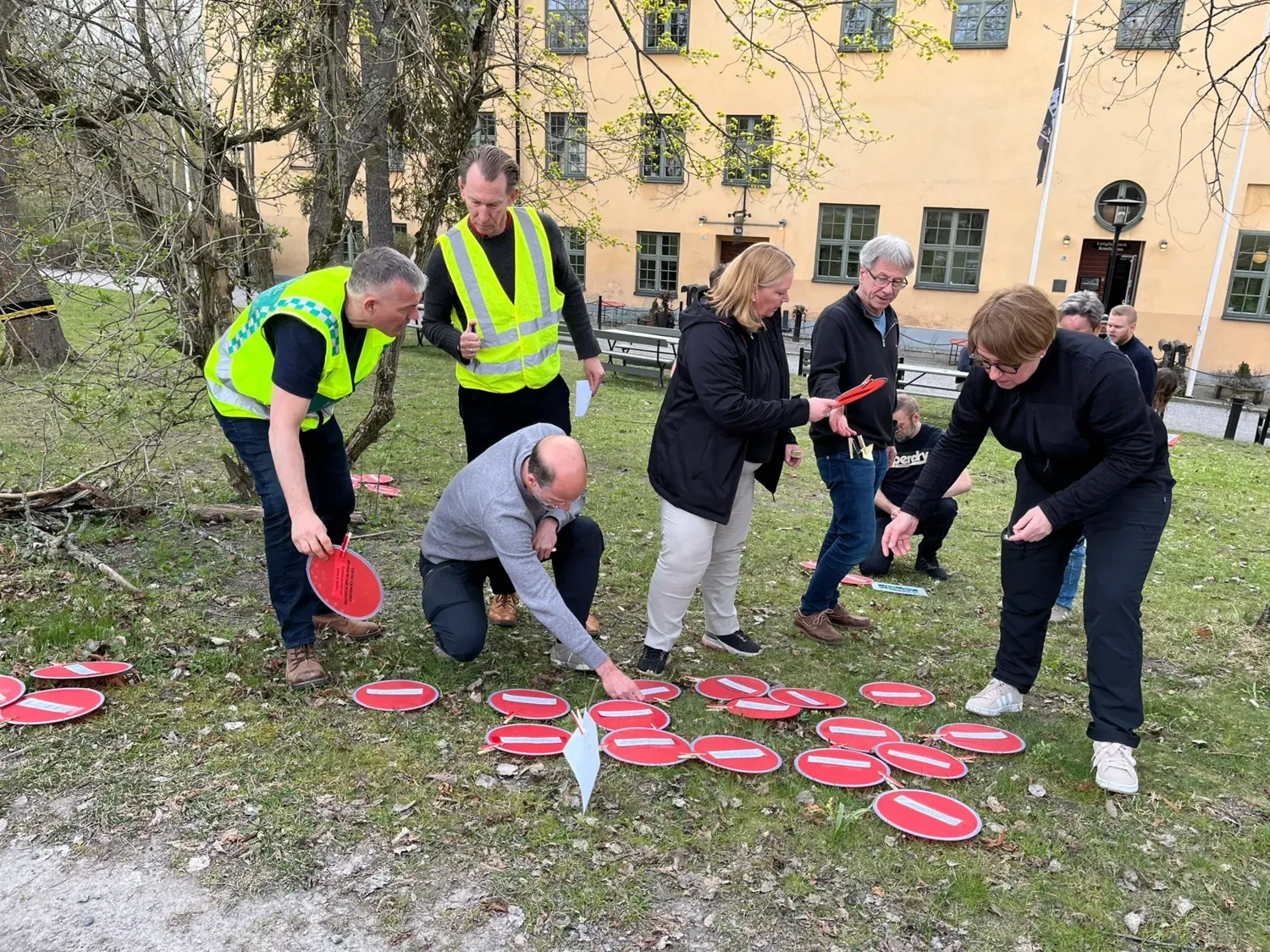 Several people, two of whom are wearing high-visibility vests, are standing outdoors on a grassy area, sorting red Frisbee-like discs. A yellow building can be seen in the background