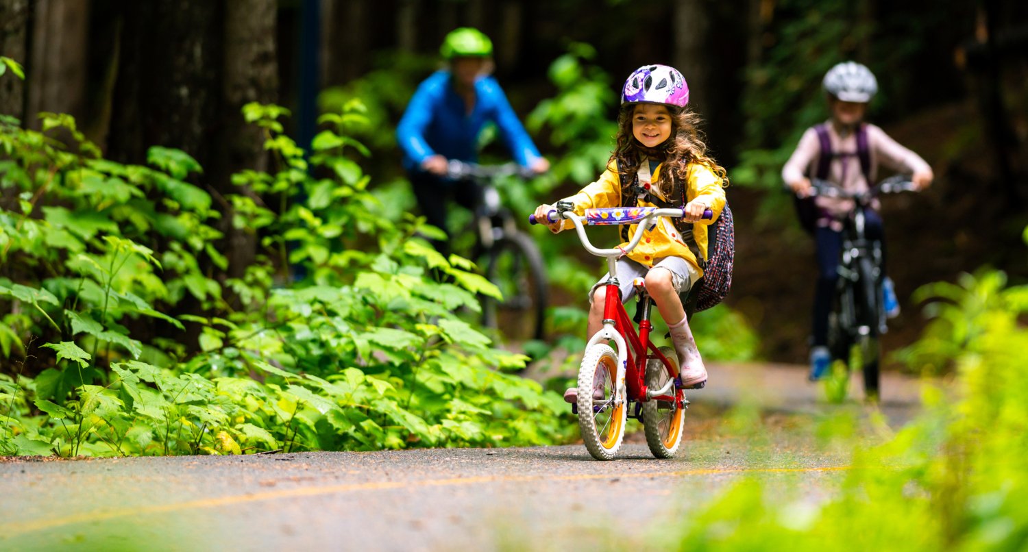 A little girl woth helmet on her bike in a forest. In the background her mother and another child.