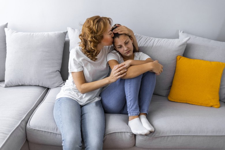 Mother holding her sad teenage daughter on a white couch.