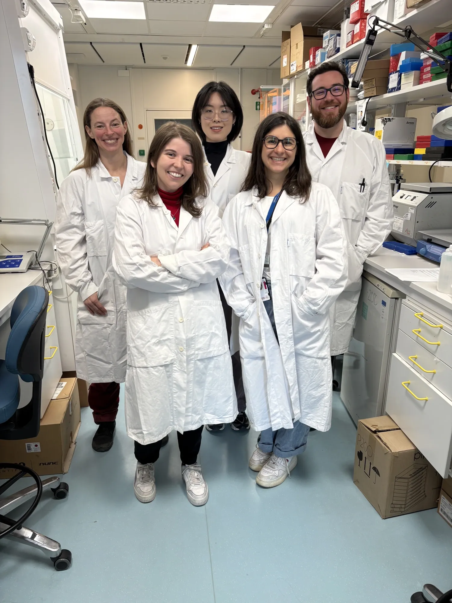 A group photo of five people. They are standing in a laboratory and are wearing white lab coats. They are smiling at the camera.