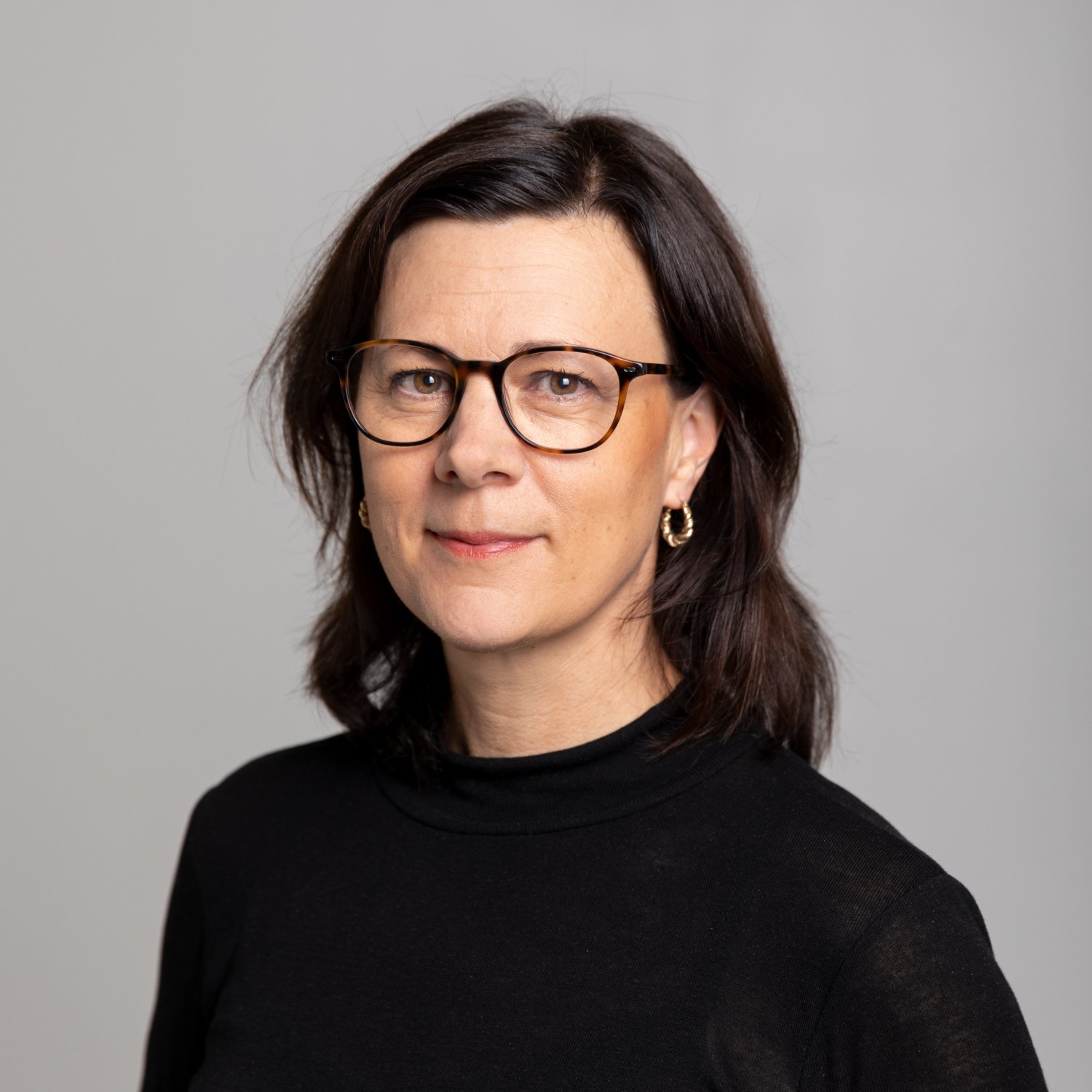 A portrait of Monica Hultcrantz. She wears a black shirt and is standing in front of a gray back drop.
