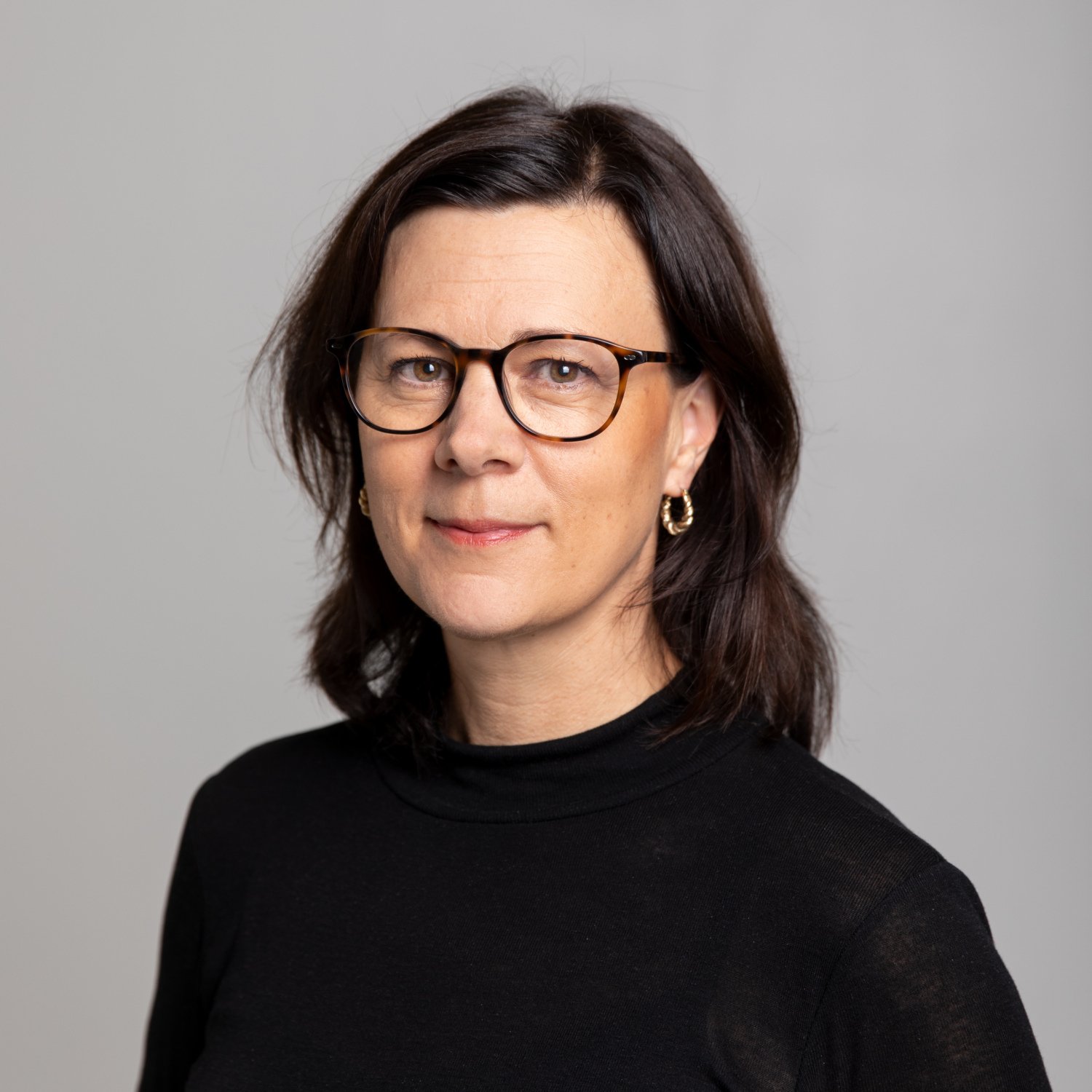 Porträtt av Monica Hultcrantz. Hon bär en svart tröja och står mot en grå bakgrund.