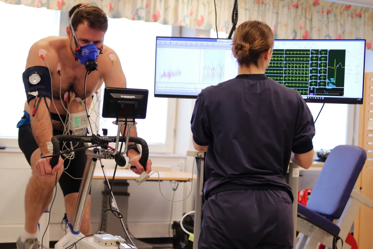 A person does a work test on a test bike. The person has a breathing mask for gas analysis and ECG electrodes attached to them. A test supervisor monitors the test and looks at a screen showing different curves of the test.