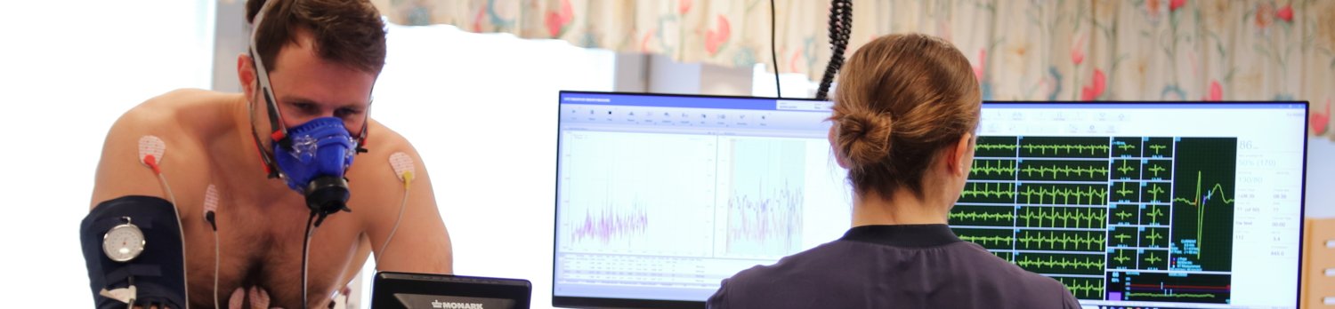 A person does a work test on a test bike. The person has a breathing mask for gas analysis and ECG electrodes attached to them. A test supervisor monitors the test and looks at a screen showing different curves of the test.
