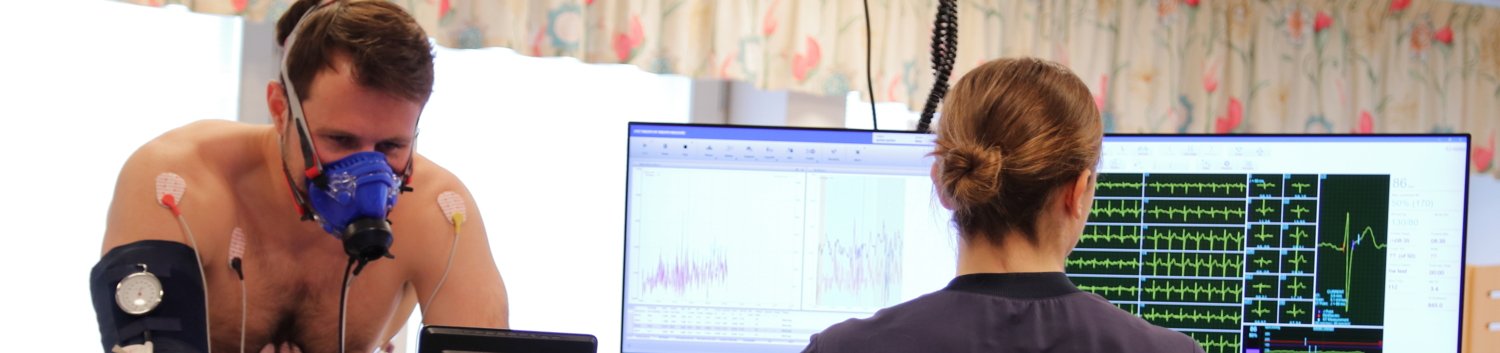 Bilden visar en testperson som gör arbetsprov på en testcykel. Personen har en andningsmask för gasanalys och EKG-elektroder kopplade till sig. En testledare övervakar testet och tittar på en skärm som visar olika kurvor över testet.