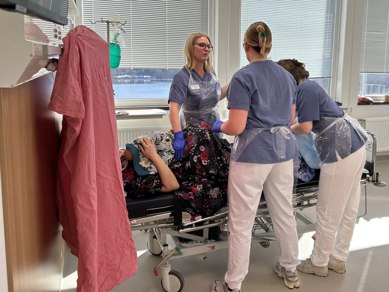 Three people in scrubs are standing around a bed on which lies a person holding a doll