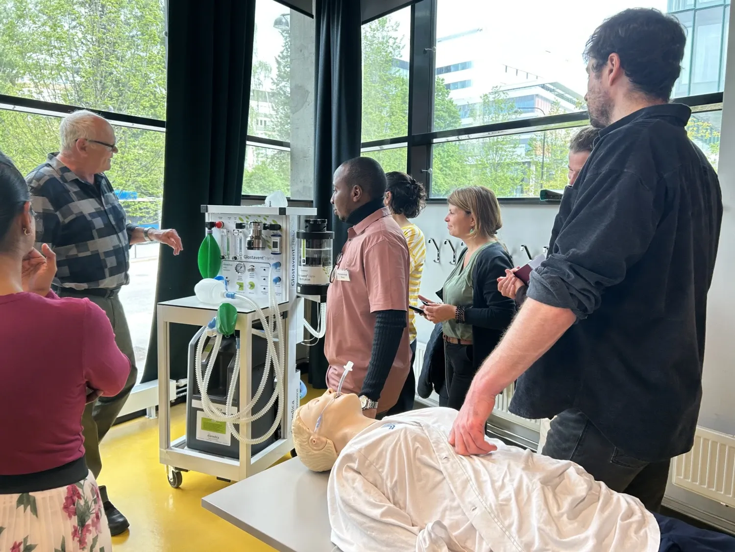 A group of people are standing around a machine, and a first aid manikin is lying on a table
