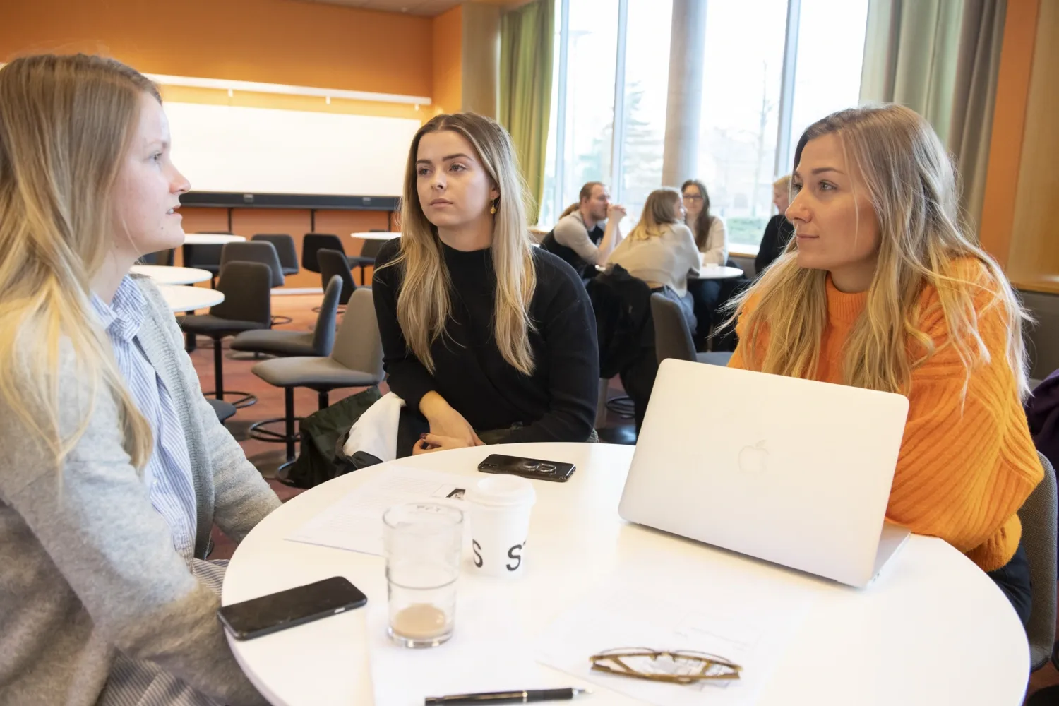 3 studenter vid sjuksköterskeprogrammet sitter runt ett bord i en föreläsningssal och diskuterar. Studenter vid ett annat bord syns i bakgrunden.