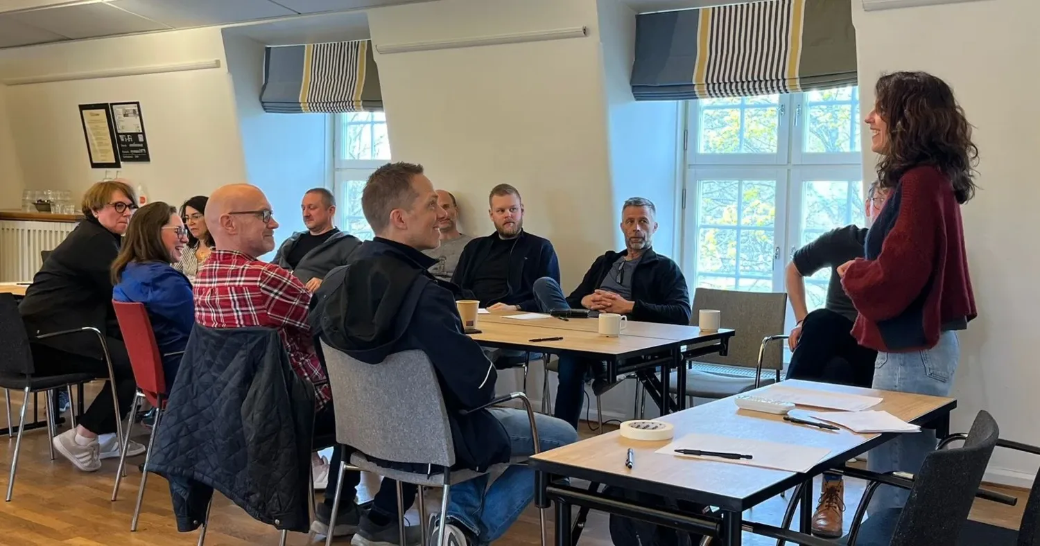 Ett bord med papper, pennor och kaffekoppar, och människor som sitter runt det. En kvinna står vid ena kortsidan av bordet