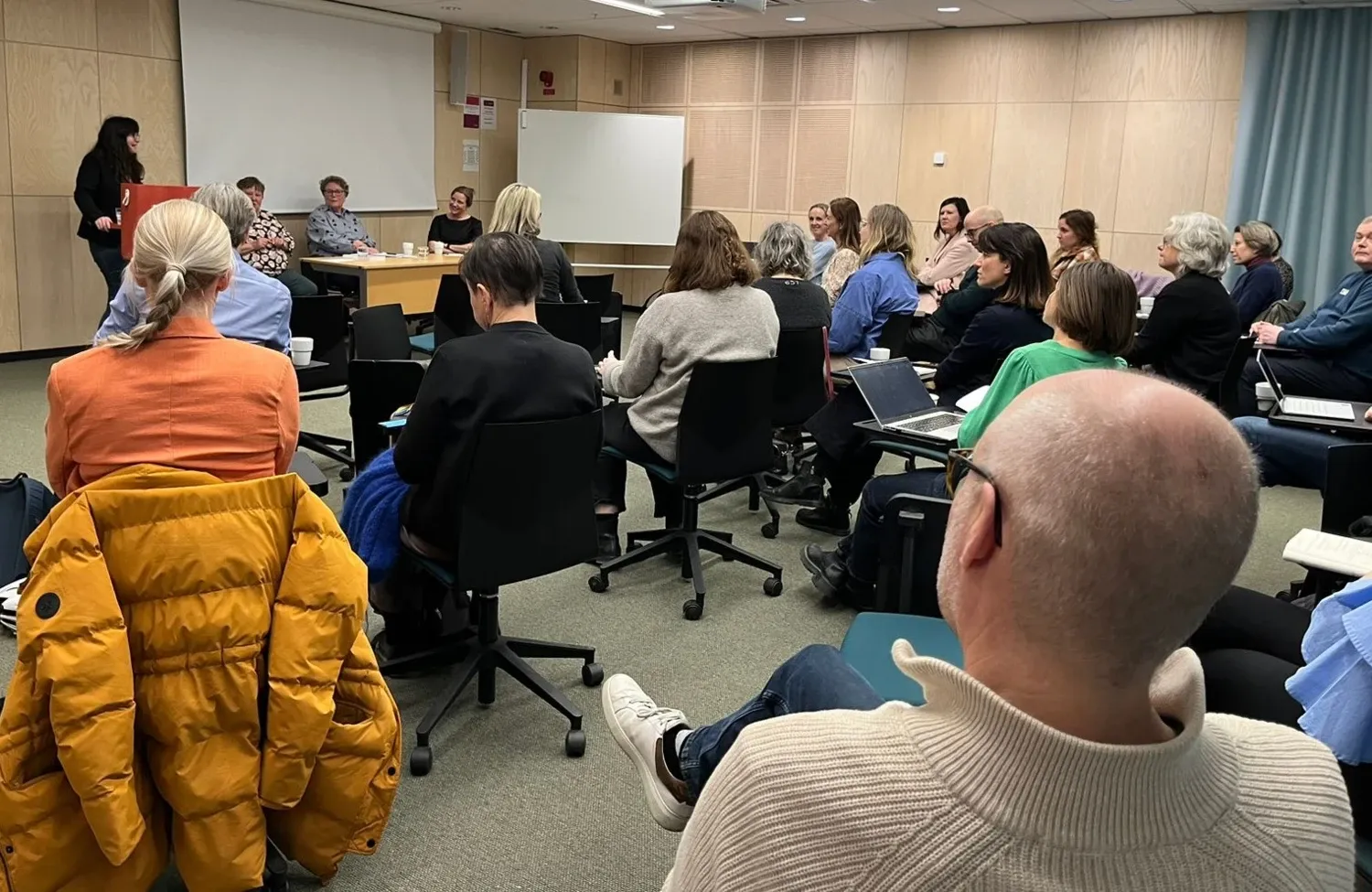 En grupp människor sitter med ryggen mot kameran och tittar på en panel bestående av tre kvinnor och en moderator