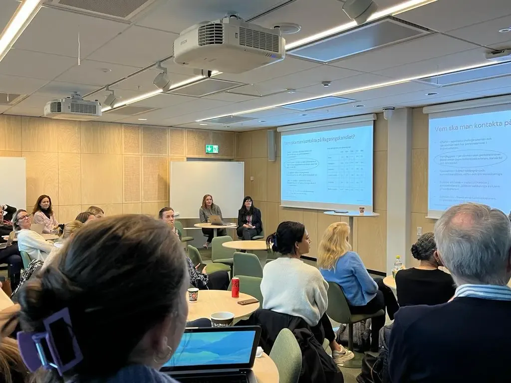A room with people sitting around small tables looking up front at a screen with a powerpoint presentation on