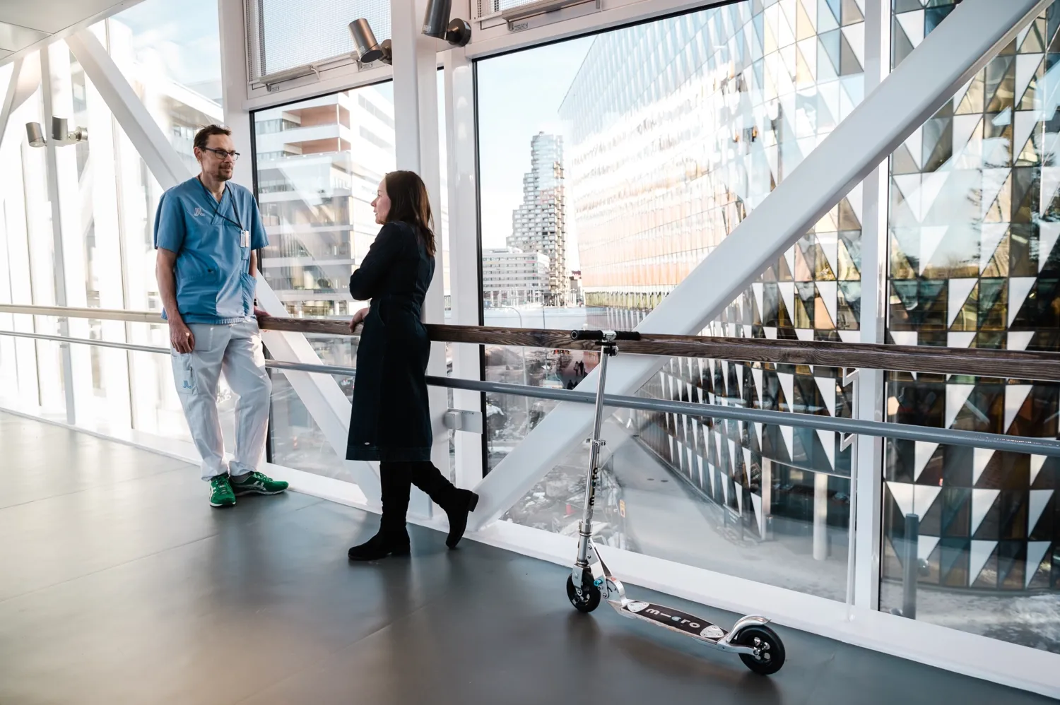 Man in hospital scrubs working at Karolinska University Hospital talks to woman in black clothes. They are standing in the corridor that leads between KI and the hospital.