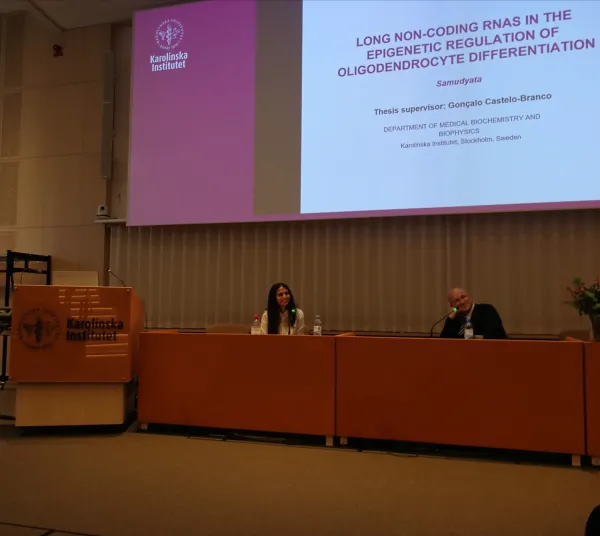 Samudyata sitting in a lecture hall in front of a screen with presentation, together with Igor Ulitsky.