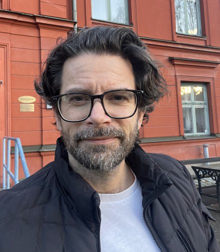 Ansiktporträtt av Gergö Hadlaczky som står utomhus framför en röd byggnad. Han har en glasögon, en vit tshirt och en svart jacka.