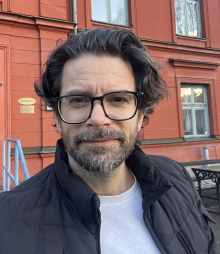 Portrait of Gergö Hadlaczky. He is standing outdoors in front of a red building. He is wearing glasses, a white tshirt and a black jacket.
