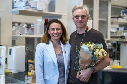A woman and a man stand next to each other and smile at the camera. The man is holding a bouquet of flowers.