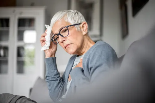 An elderly worried woman in glasses sits and holds her chest.