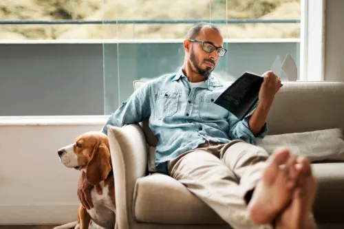 A man in his forties is sitting on a sofa reading a book. He is relaxed and has put his feet up on the table.