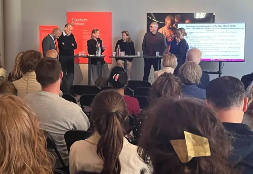 Six people are standing behind a high table, with an audience seated in front of them.