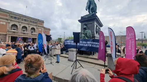 A number of people have gathered in the square. There is a stage where Caroline reads out testimonials. On the stage there are banners showing who the organizers are.