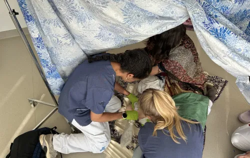 Two course participants dressed in hospital gowns practise providing care on the floor of a tent.